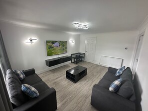 Un salon moderne avec un téléviseur à écran plat fixé au mur, un meuble de rangement noir en dessous, deux canapés noirs avec des coussins à carreaux, une table basse et un plancher en bois.