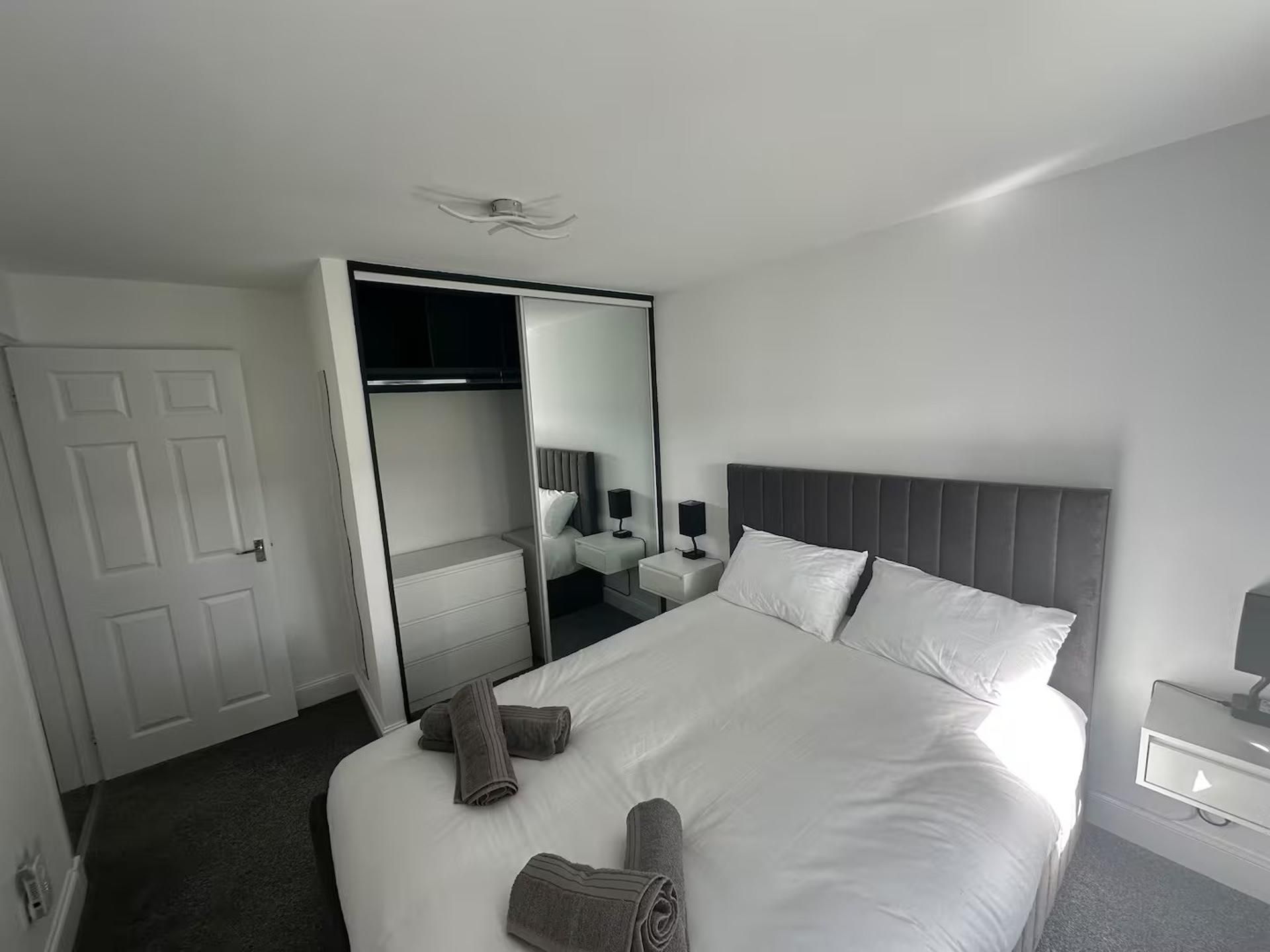 A modern hotel room with a large bed, a bedside table, a nightstand, a mirror, and a wardrobe.