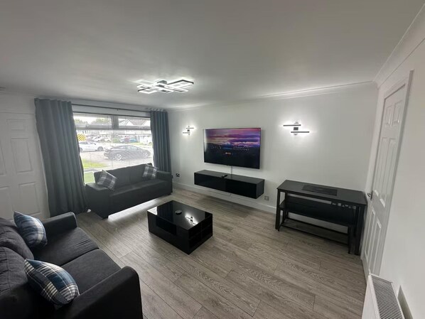 Un salon moderne avec un canapé, une table basse, un téléviseur fixé au mur et une fenêtre avec des rideaux.
