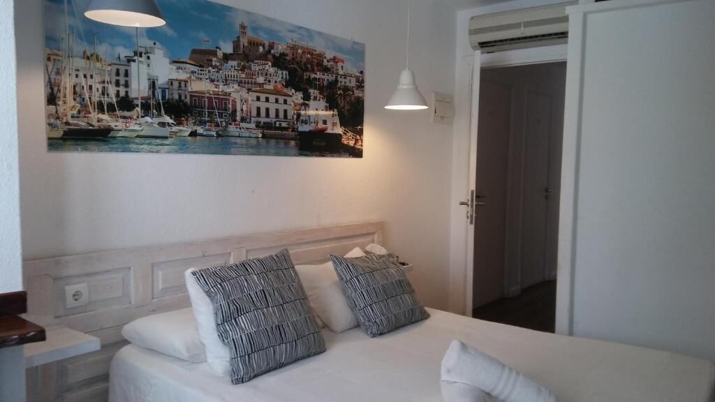 Habitación de hotel con cama, ropa de cama blanca, cabecero, un cuadro de un pueblo costero y una puerta.