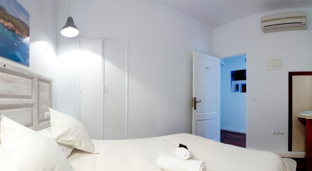 Una habitación de hotel con cama, puerta, unidad de aire acondicionado y baño visible a través de una puerta abierta.