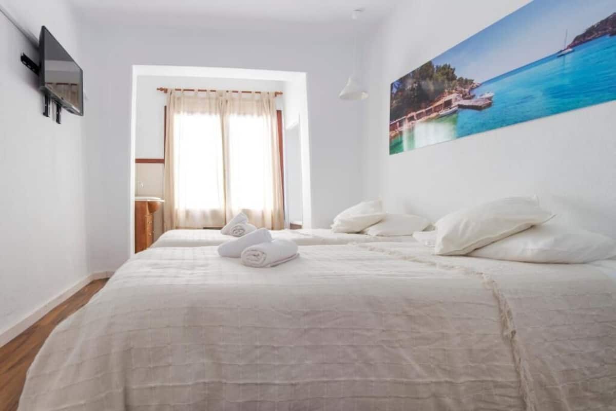 A bedroom with a large bed, a painting of a coastal scene, and a window with curtains.
