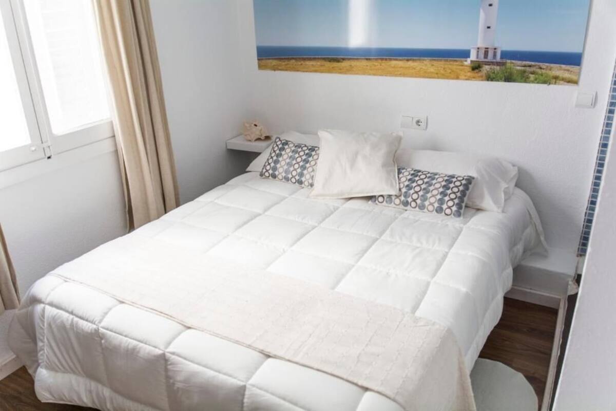 A neatly made bed with a white duvet and patterned pillows, a small bedside table with a lamp, and a painting of a lighthouse on the wall.