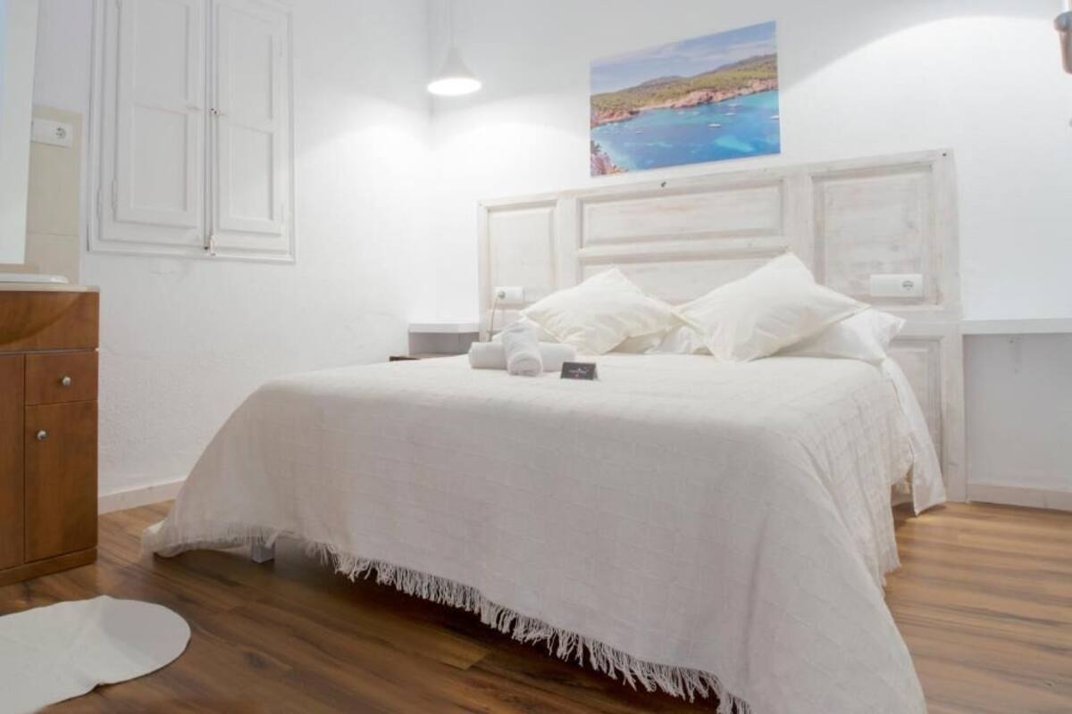 A bedroom with a white bed, wooden floor, and a painting of a coastal scene on the wall.