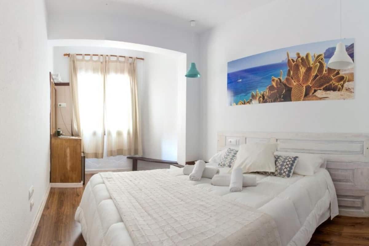 A bedroom with a large bed, a nightstand, a painting of a beach scene, and a window with curtains.