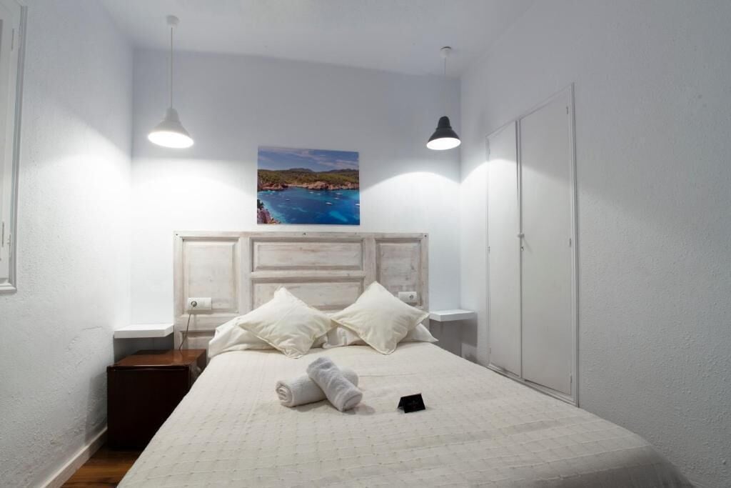 Un lit bien fait, recouvert d’une courtepointe blanche, avec deux oreillers et une serviette enroulée posée sur le sol. On y trouve une petite table de chevet avec une lampe, un tableau représentant un paysage côtier accroché au mur, et une armoire avec un miroir.