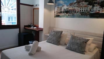 Une chambre d’hôtel avec un lit, un tableau représentant une ville côtière, une fenêtre donnant sur des immeubles et un lavabo avec un miroir.