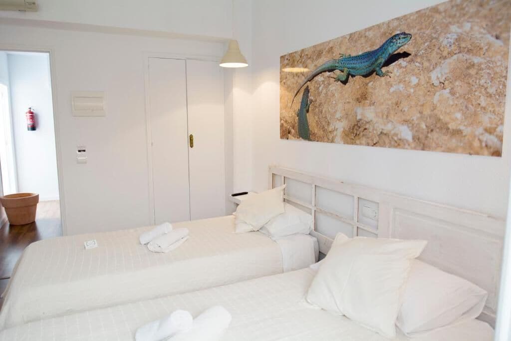 Quarto de hotel com duas camas, roupa de cama branca, um quadro de um lagarto na parede e uma porta com extintor de incêndio.