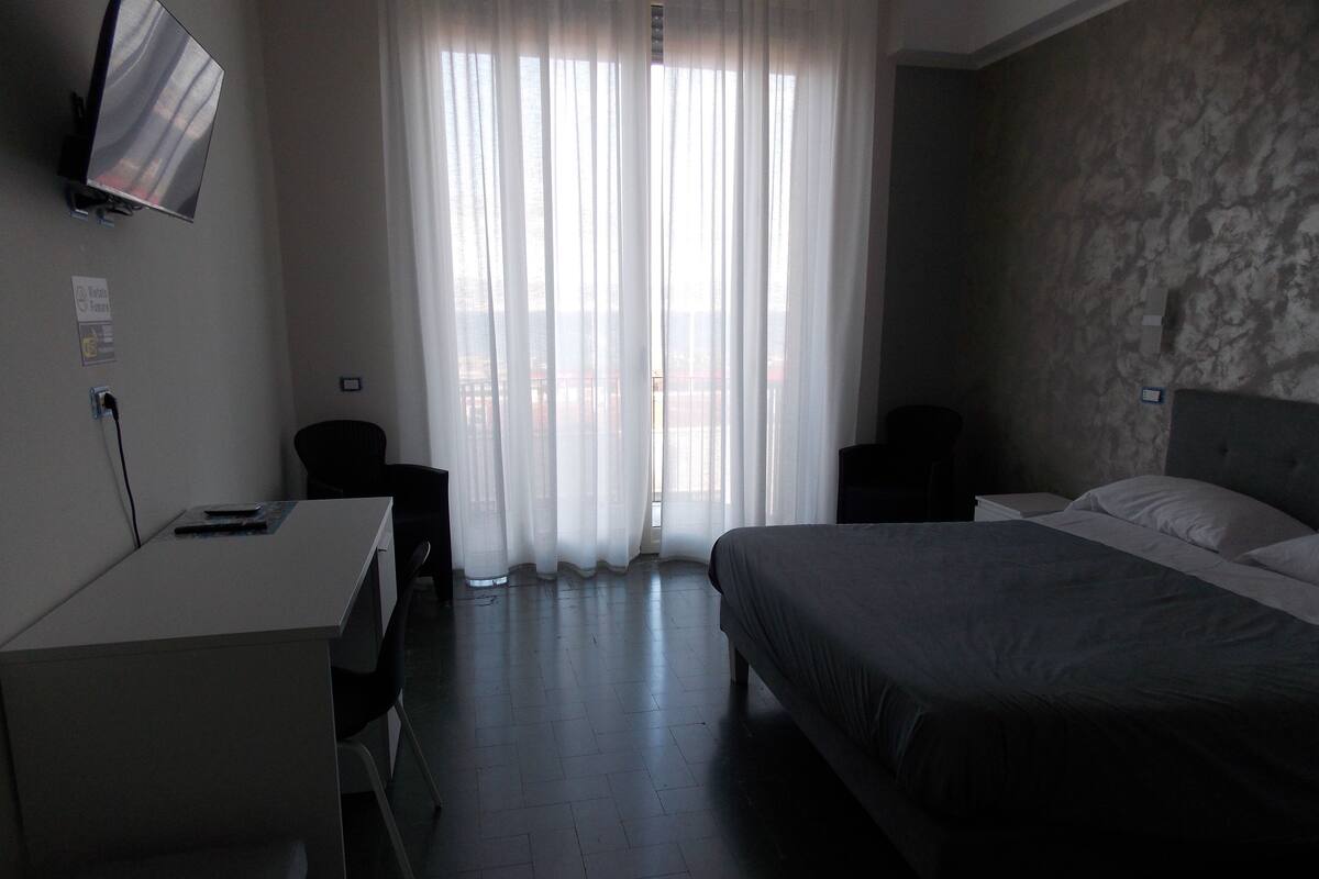 A hotel room with a bed, a desk, a chair, a TV, and a window with sheer curtains.