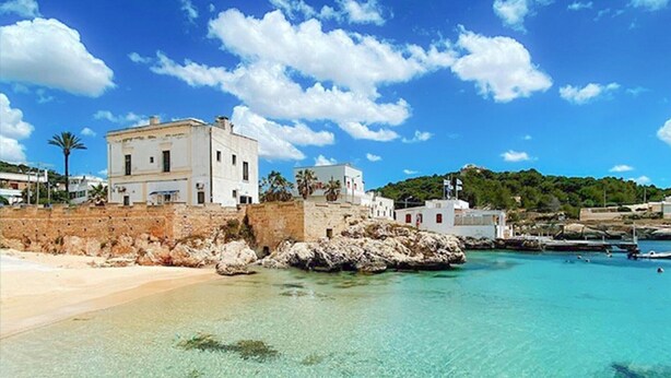 Un edificio costero con fachada blanca, situado cerca de un mar azul cristalino y una playa de arena.