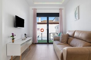 Ein modernes Wohnzimmer mit einer Couch, einem Flachbildfernseher, einem weißen Konsolentisch mit Pflanze und einer Glastür, die zu einem Außenbereich führt.