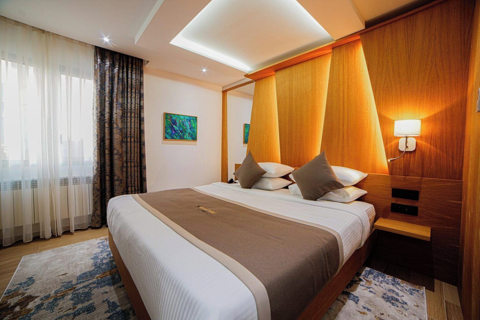 Habitación de hotel con una cama grande, cabecera de madera, mesita de noche, lámpara y un cuadro en la pared.