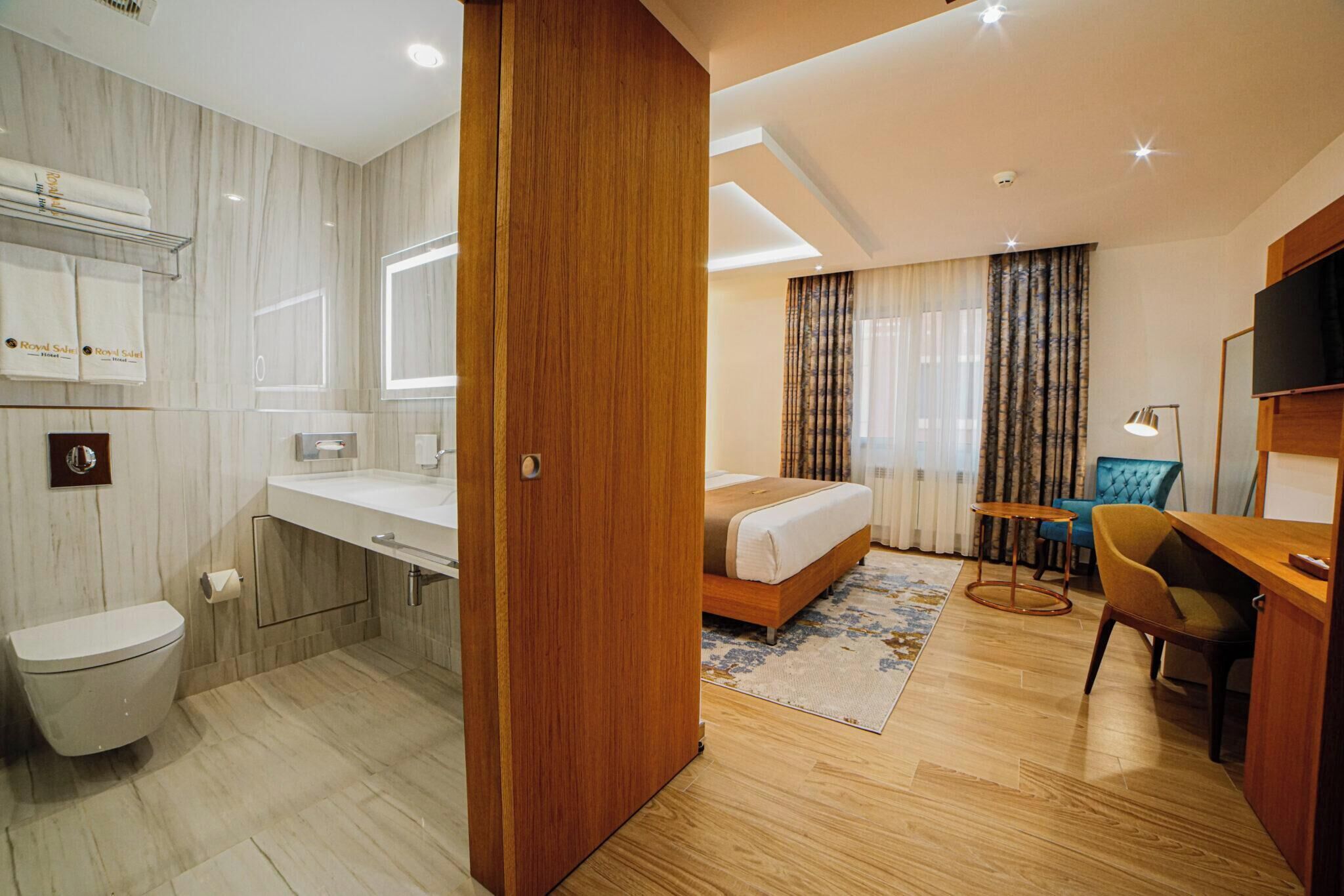A hotel room with a wooden door, a bed, a desk, a chair, a TV, a bathroom with a toilet, a sink, and a shower.