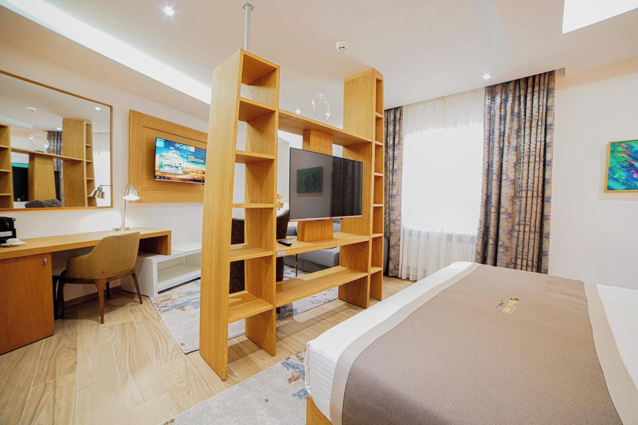 A modern hotel room with a wooden desk, a television, a bed, and a large window with curtains.