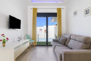 Ein modernes Wohnzimmer mit Flachbildfernseher, einer Couch, einem kleinen Tisch mit Dekoration, einem Sessel und einer Schiebeglastür, die zu einem Außenbereich führt.