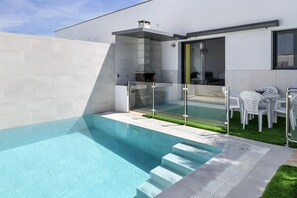 Ein moderner Poolbereich im Freien mit einem gläsernen Whirlpool, einem Grill und einer Sitzecke mit weißen Kunststoffstühlen.
