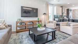 En moderne stue med et fladskærms-tv, to sofaer, et sofabord og et køkken i baggrunden.