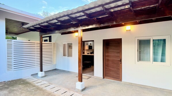 Un espacio al aire libre techado con una pérgola de madera, una pared blanca con una puerta y una pequeña cocina visible a través de otra puerta abierta.
