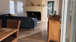 Een ruime woonkamer met een open haard, een bank, een eettafel met stoelen en uitzicht op de omgeving door een deur.
