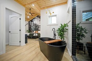 Un baño moderno con una bañera independiente, suelo de madera, ventilador de techo y un amplio ventanal.
