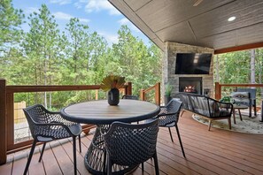 Eine überdachte Terrasse im Freien mit einem runden Tisch und Stühlen, einem Kamin aus Stein und einem an der Wand montierten Fernseher.