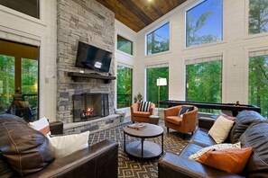 A spacious living room with a stone fireplace, leather sofas, and large windows offering a view of the outdoors. - 2 King Suites, Outdoor Fireplace, Games, Hot Tub (Broken Bow)