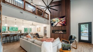 A spacious living area with a high ceiling, a large TV mounted on the wall, a fireplace, and a balcony with a wooden deck. - StayHocha Luxury Pet Friendly Cabin (Broken Bow)