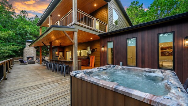 A wooden deck with a hot tub, outdoor seating, and a stone fireplace. - StayHocha Luxury Pet Friendly Cabin (Broken Bow)