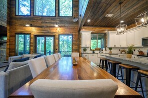 A spacious dining area with a long wooden table and multiple chairs, a kitchen with white cabinets and a large window, and a view of the surrounding forest. - Luxurious 4 Bedroom Cabin Retreat in Broken Bow (Broken Bow)