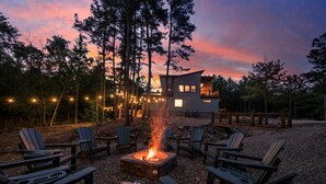 Ett modernt utomhusområde med sittplatser, Adirondack-stolar och en eldstad, omgivet av träd och ett trädäck.