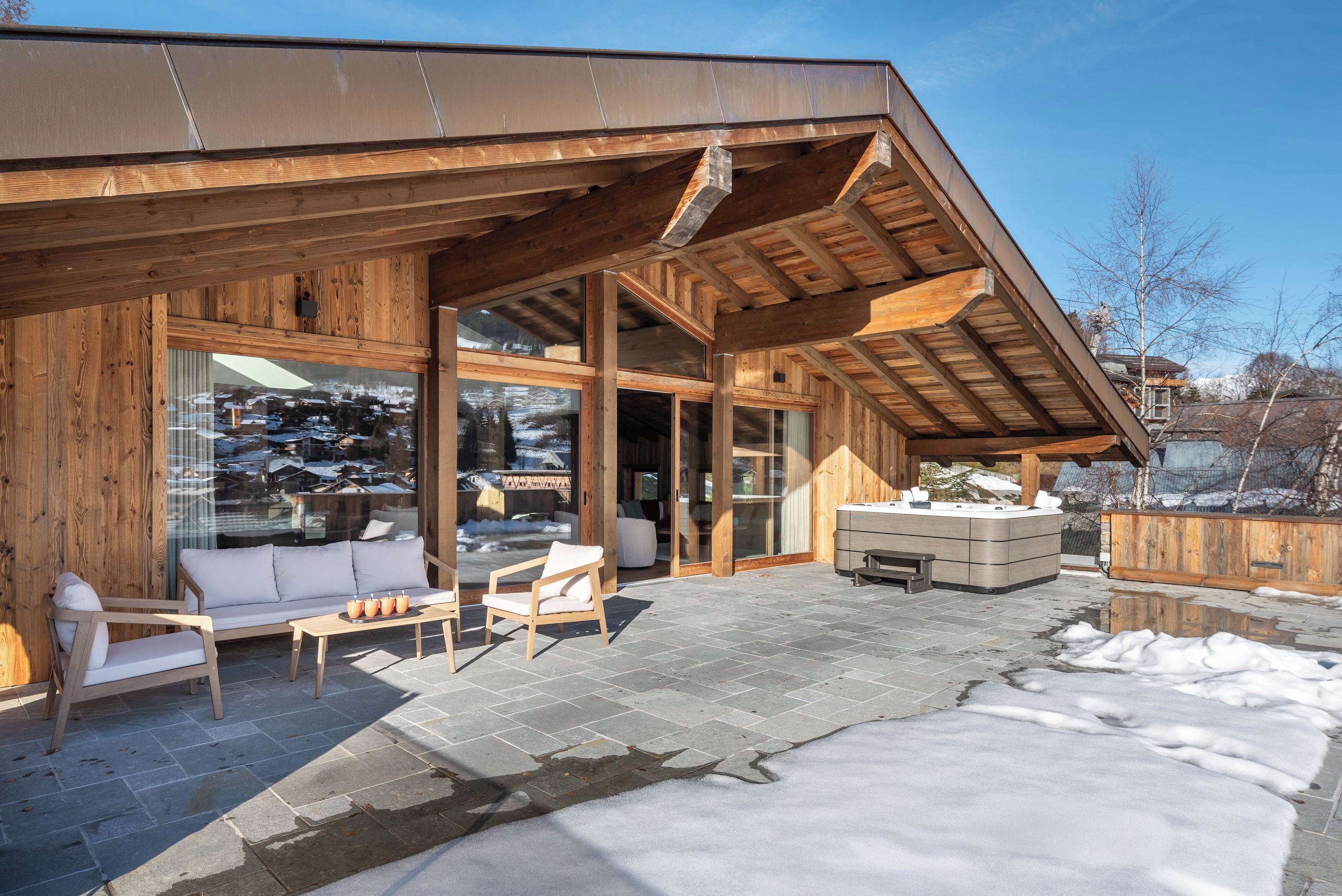 A wooden cabin with a stone patio, a sofa, chairs, and a hot tub.