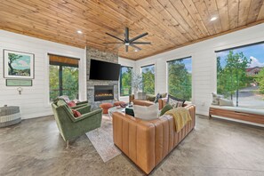 A spacious living room with a fireplace, large windows, and a ceiling fan. - Magical Spacious cabin on flowing creek for Groups (Broken Bow)
