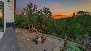 Un patio con fogata y mobiliario exterior, rodeado de árboles y vistas al atardecer.