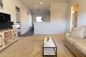 Una sala de estar con televisor, una mesa de centro con flores, un sofá beige y una cocina al fondo.