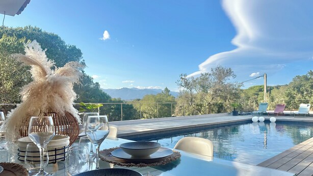 Mesa de comedor lista con platos, tazones y vasos, con vista a una piscina y montañas.