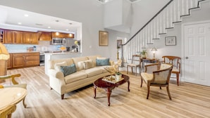 A spacious living area with a sofa, coffee table, and chairs, a staircase, and a kitchen in the background. - 5BR Estate • Hot Tub & Sleeps 10 • Near Beach (Edgewater)