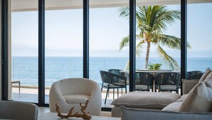 Uma sala de estar moderna com uma grande janela com vista para o oceano, mobiliada com um sofá, cadeiras e uma mesa.