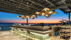 Área de bar ao ar livre com vista para o pôr do sol, um balcão, banquetas e um carrinho.