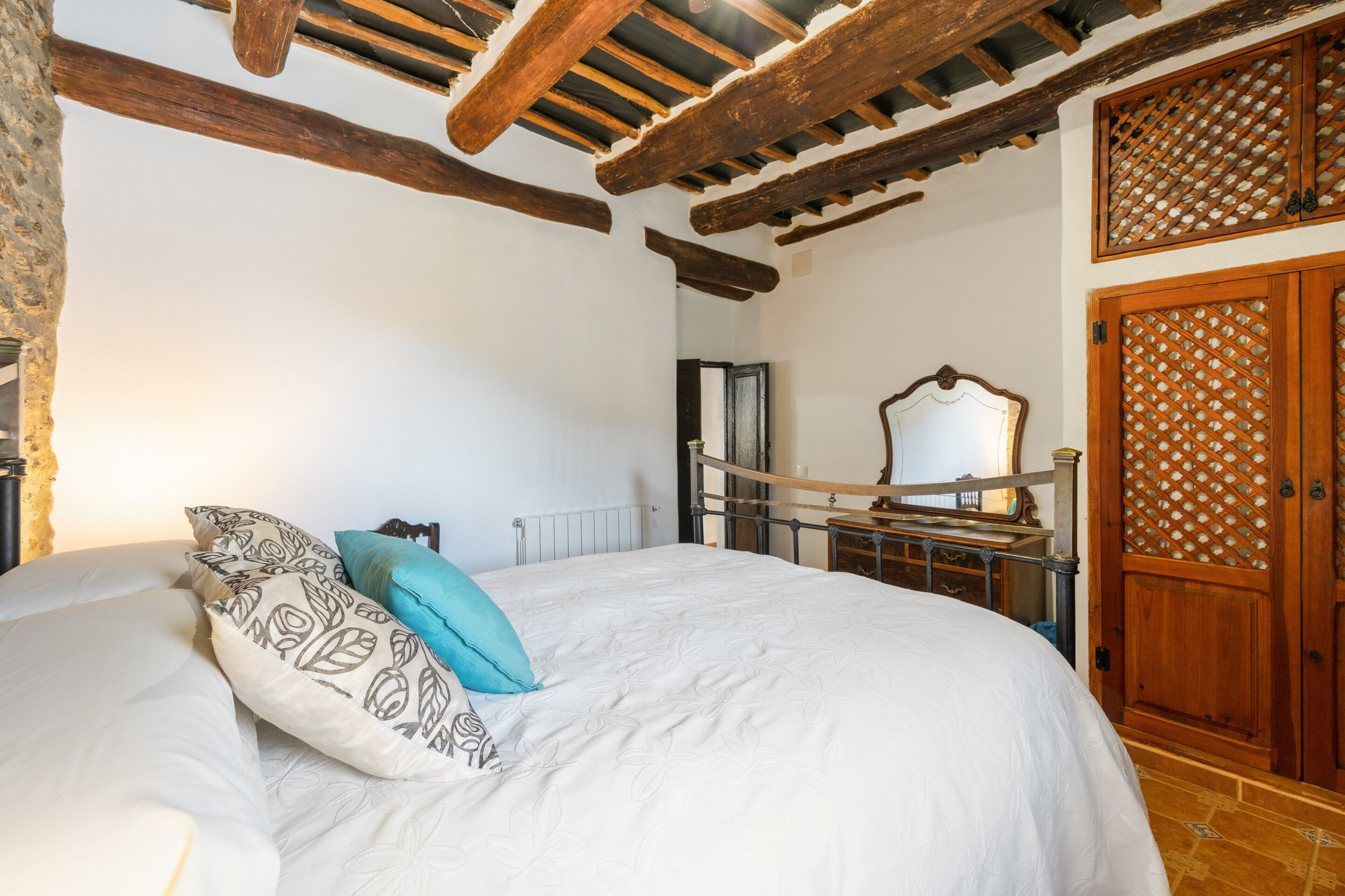 Un dormitorio con una cama, vigas de techo de madera, una puerta de madera con diseño de celosía y un espejo sobre la cama.