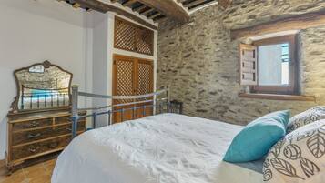 Une chambre avec un mur en pierre, un cadre de lit en bois, une commode et une fenêtre avec des volets.