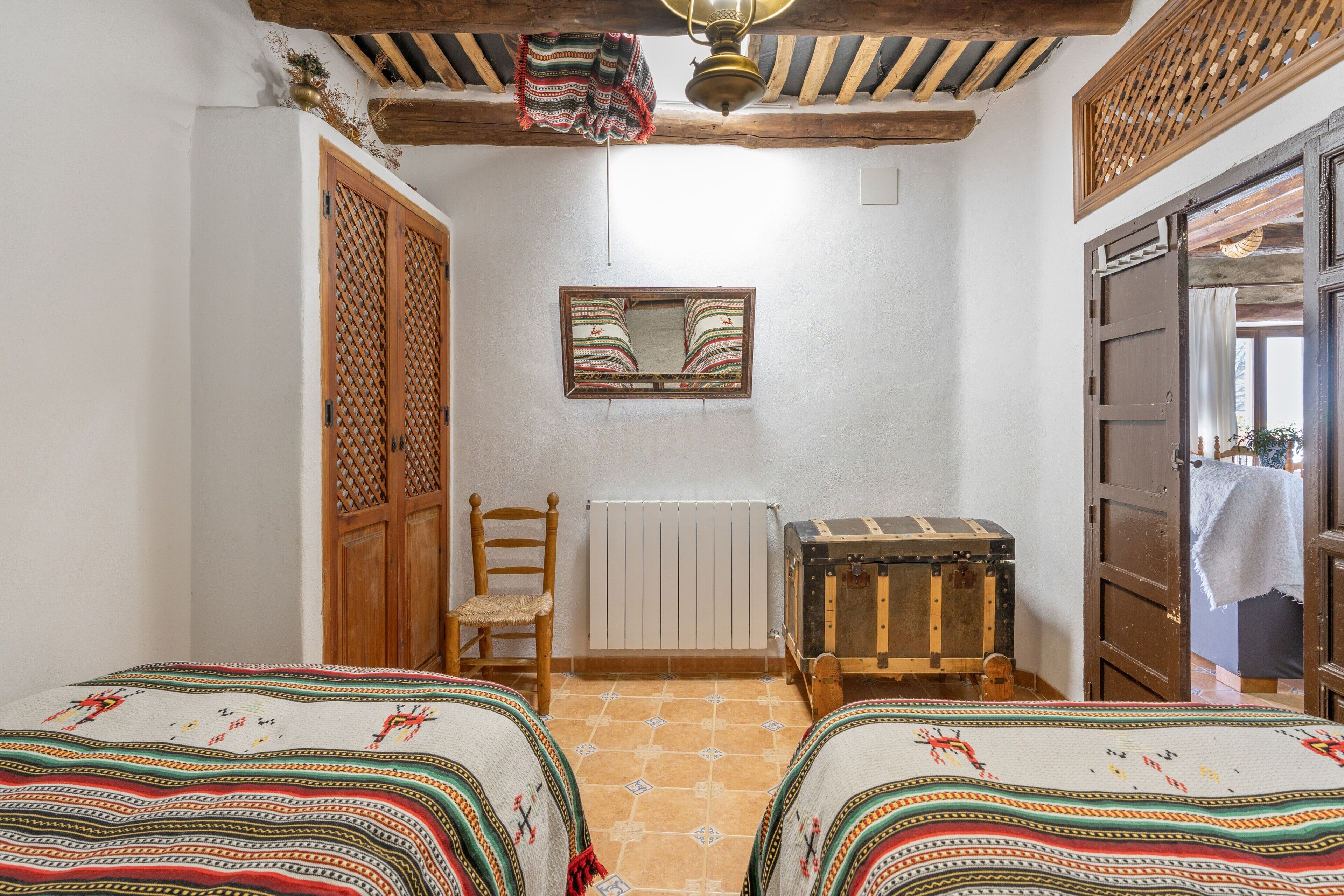 A room with two beds, a wooden chest, a radiator, and a window with a mirror.