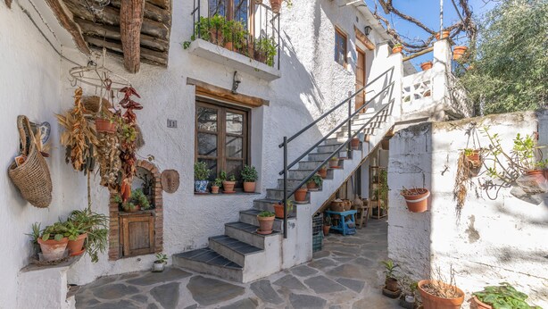 Une cour étroite, pavée de pierres, avec un escalier menant à un deuxième étage, le tout entouré de murs blancs ornés de plantes en pot et de suspensions florales.