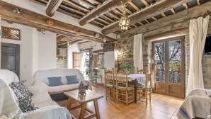 Un intérieur rustique avec un plafond en bois, un coin repas avec une table et des chaises, un canapé agrémenté de coussins à motifs et une vue dégagée sur l’extérieur grâce à une grande fenêtre.