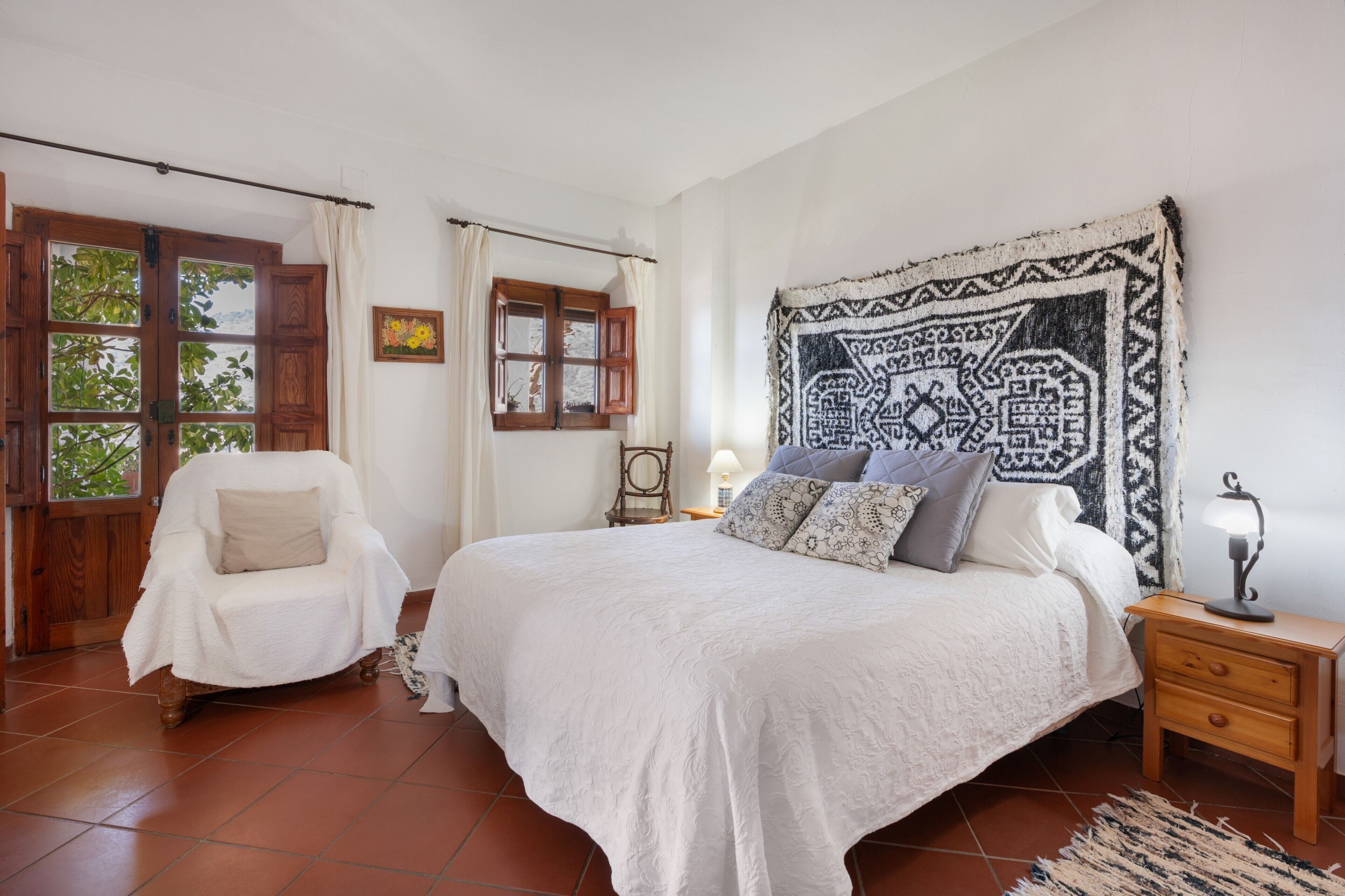 Un dormitorio con cama, una silla, una mesita de noche, una lámpara, un ventanal y un tapiz.