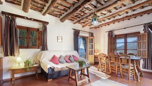 Un salón acogedor con sofá, una mesa de madera y sillas, techo de madera y ventanas grandes con cortinas.