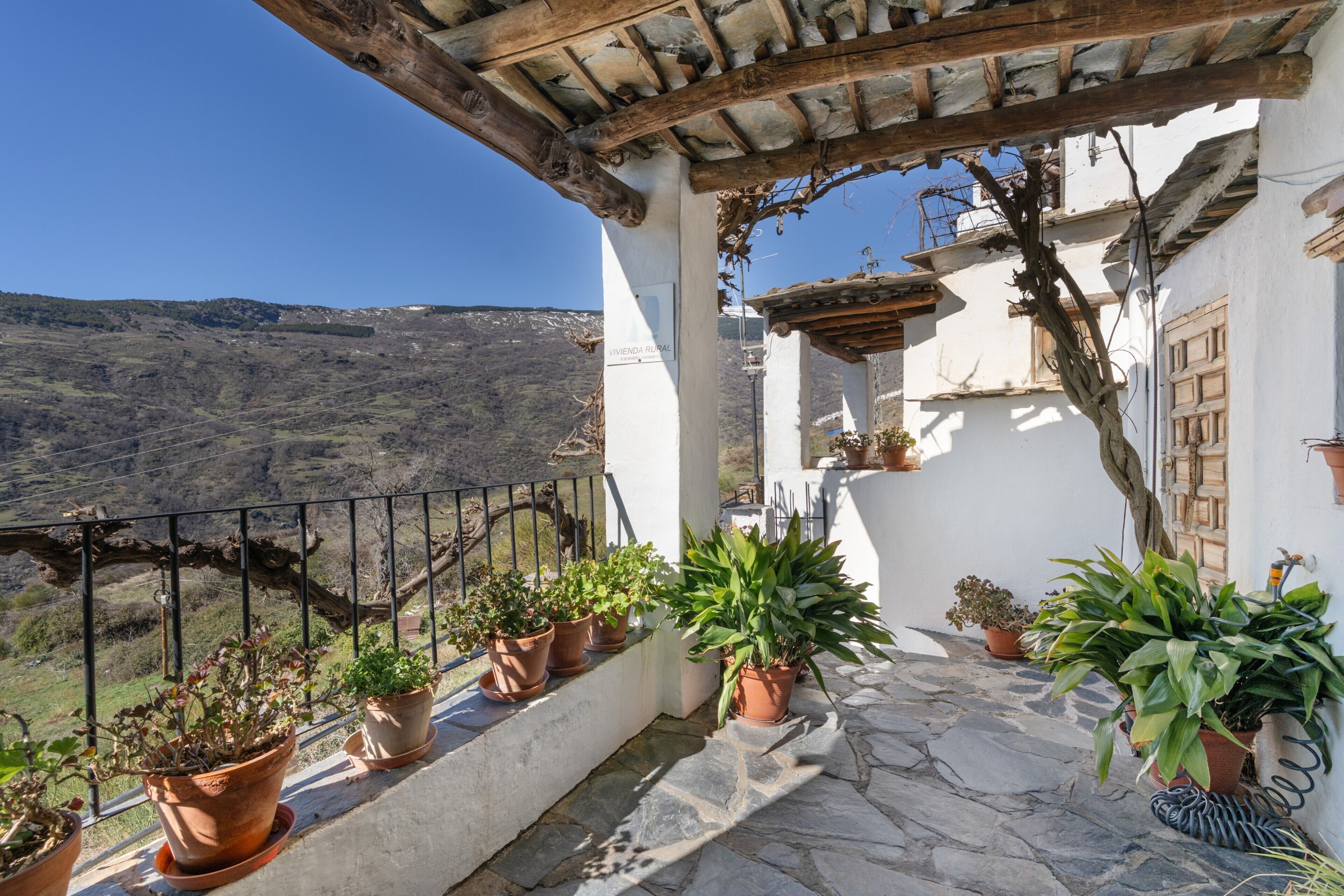 Un balcón con plantas en macetas, suelo de piedra y vistas a un paisaje montañoso.
