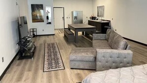 A modern living room with a grey sofa, a coffee table, and a TV on a stand. There is a kitchen area with a fridge and a microwave. The room has a wooden floor and a patterned rug. - Food Truck Court BNB w/breakfast & pool table (Carlsbad)