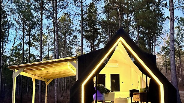 A cabin with an open front and a covered porch, surrounded by trees.