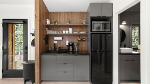 A modern kitchen with grey cabinets, a black refrigerator, and a microwave. There is a wooden wall panel and a window with a view of trees.