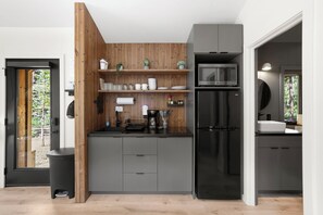 A modern kitchen with grey cabinets, a black refrigerator, and a microwave. There is a wooden wall panel and a window with a view of trees.